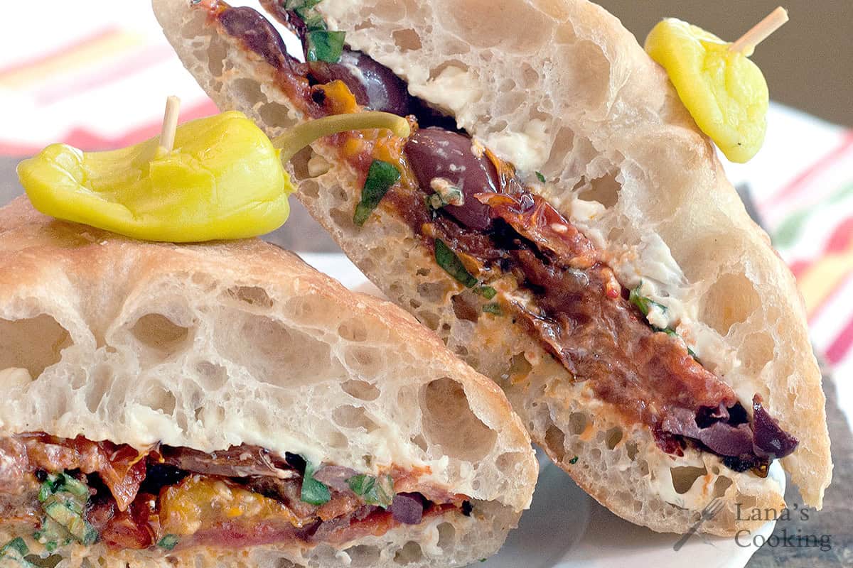 A ciabatta sandwich cut in half, filled with olives, greens, and roasted tomatoes, each half topped with a yellow pepper secured by a toothpick.