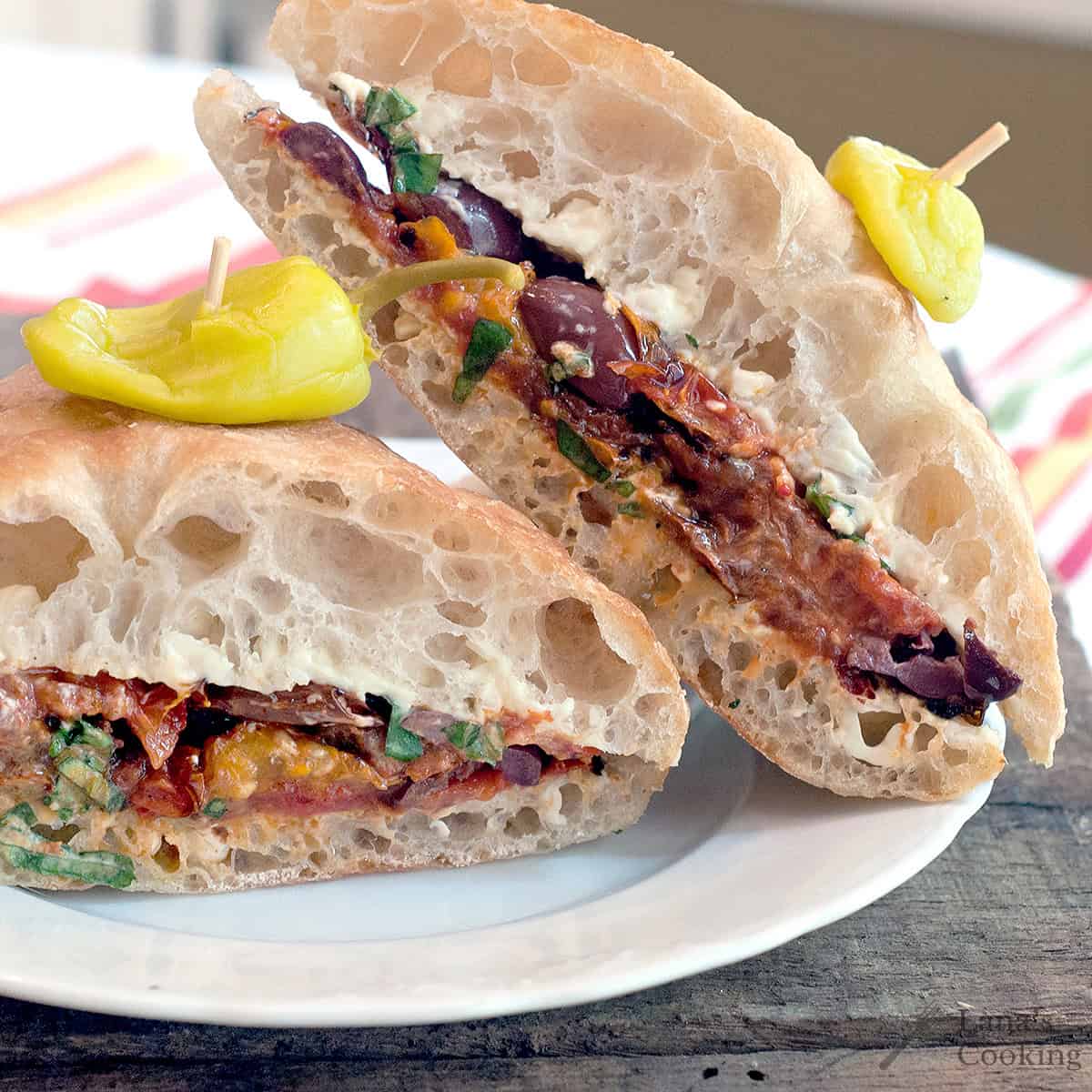 A ciabatta sandwich cut in half, filled with vegetables and cheese, served on a white plate with a yellow pepper on top.