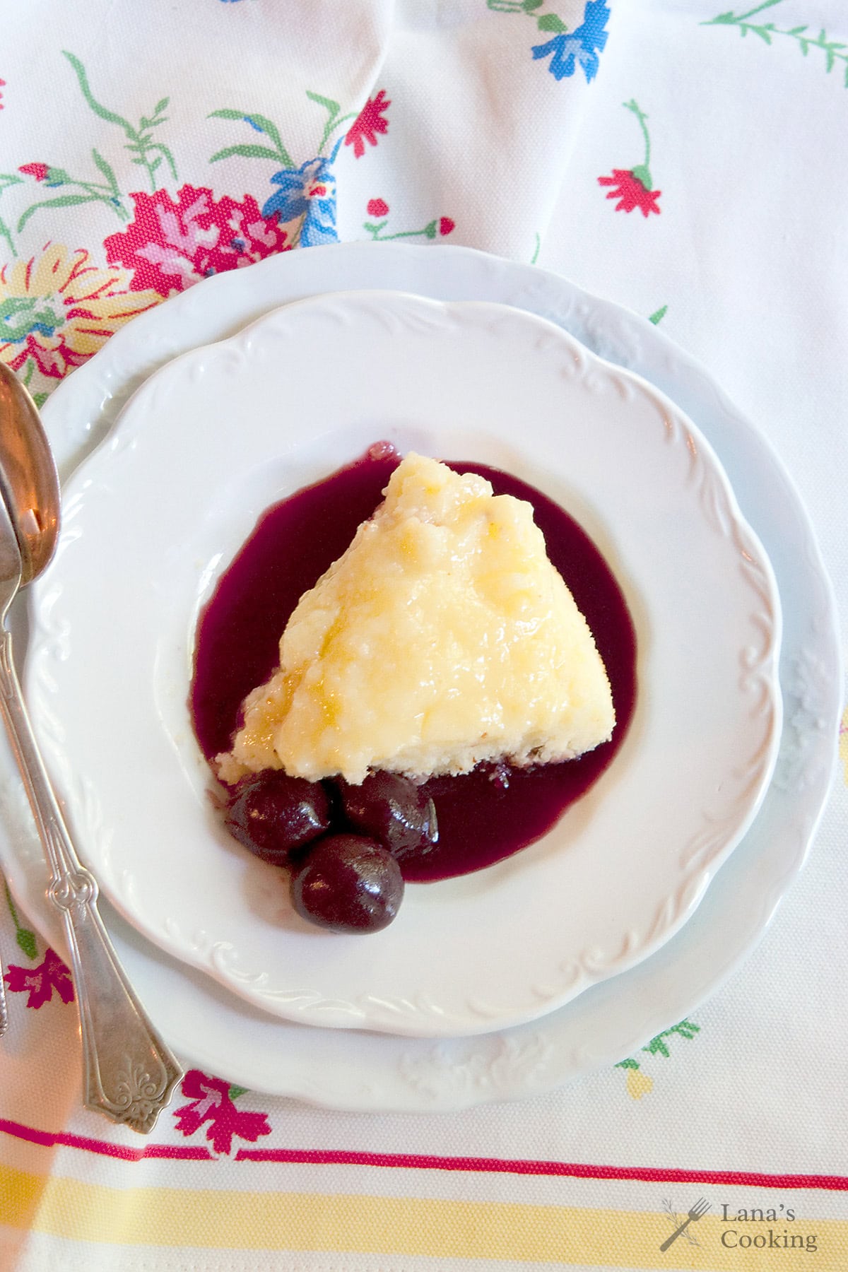 A slice of lemon pudding cake with cherry sauce and whole cherries on a white plate, with a spoon beside it.
