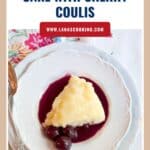 A slice of lemon pudding cake with cherry coulis and cherries on a white plate with a spoon.