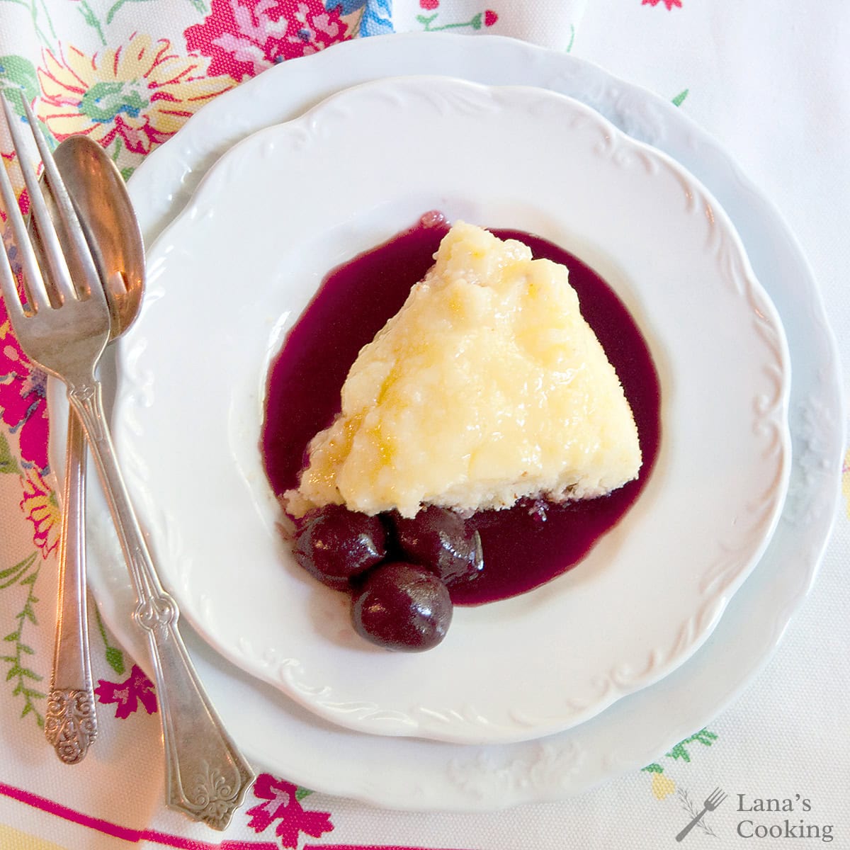 A slice of lemon pudding cake sits on a plate with dark cherry coulis and whole cherries, beside a fork and spoon on a floral tablecloth.