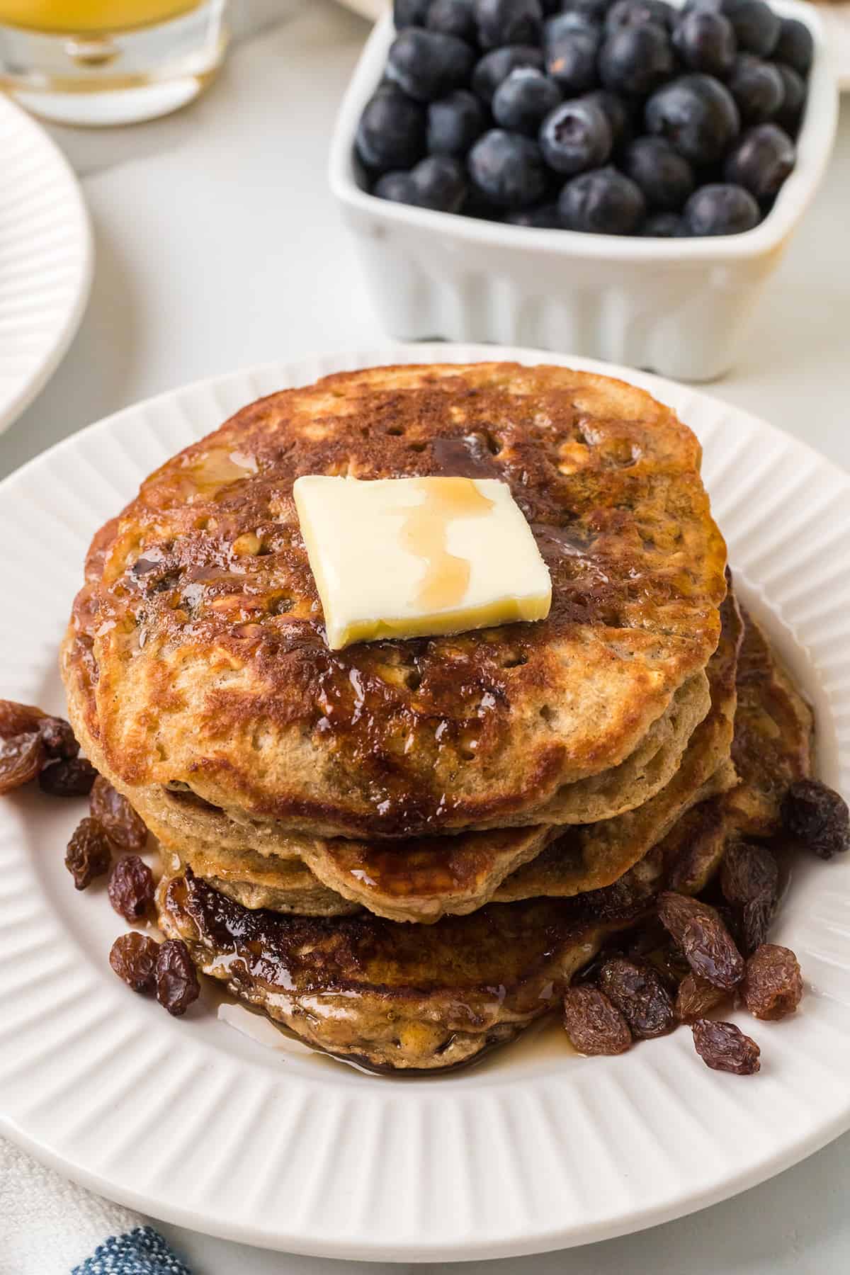 A stack of pancakes with butter on top and raisins on the side of the plate.