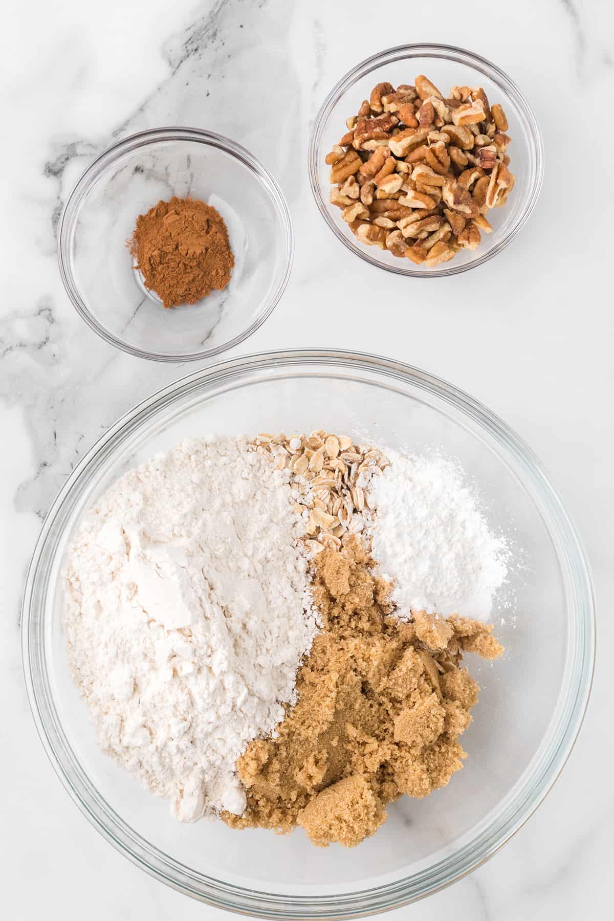 Glass bowls with flour, oats, brown sugar, cinnamon, powdered sugar, and chopped pecans on a marble surface.