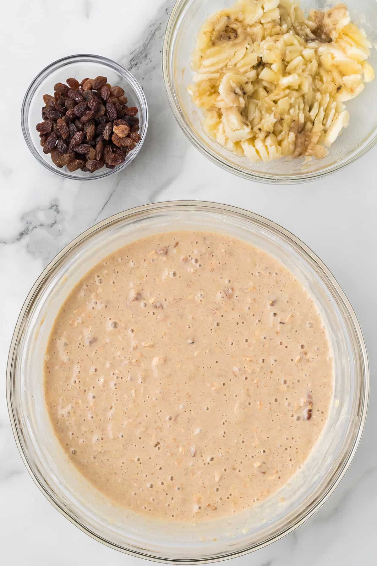 Large bowl of batter, with a small bowl of raisins and a bowl of mashed bananas on the side.