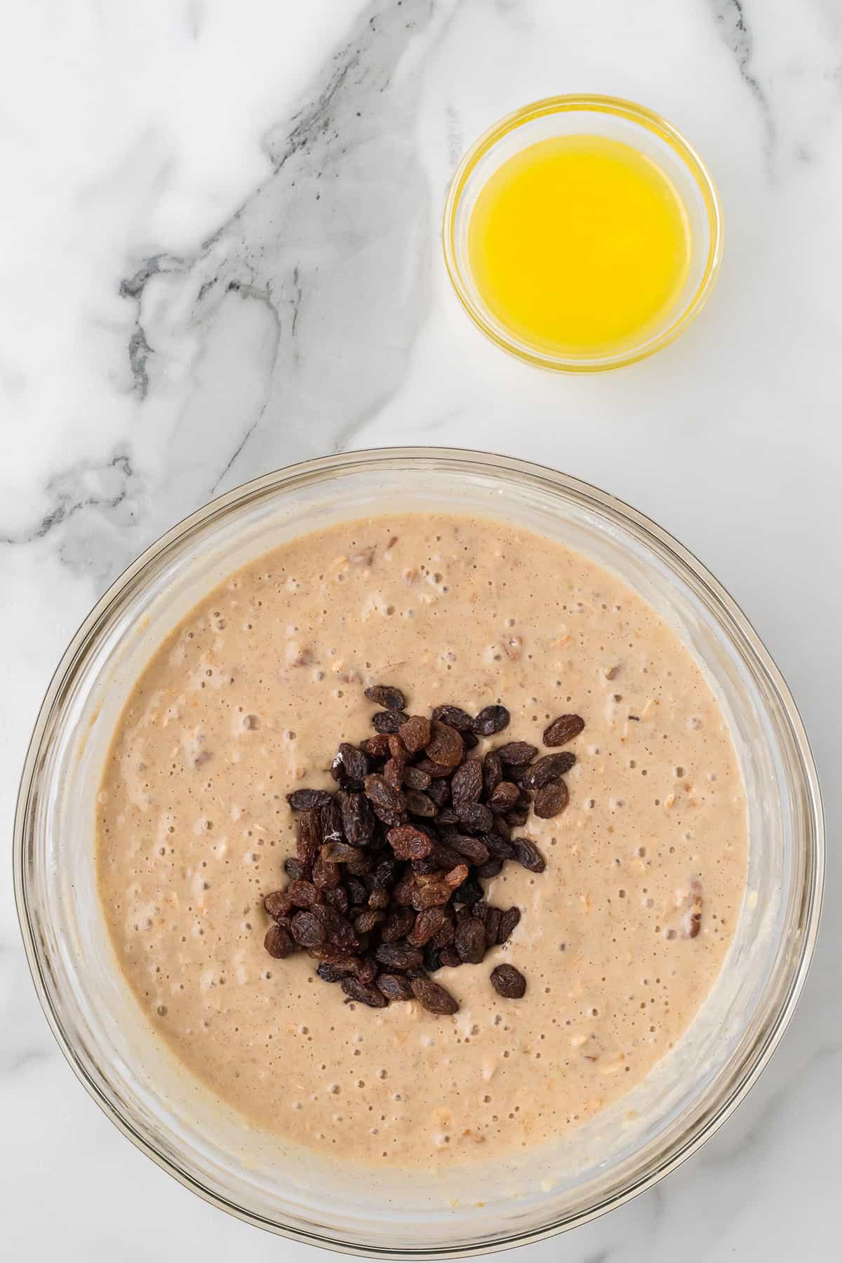 A glass bowl of batter with raisins on top, next to a small bowl of melted butter on a marble surface.