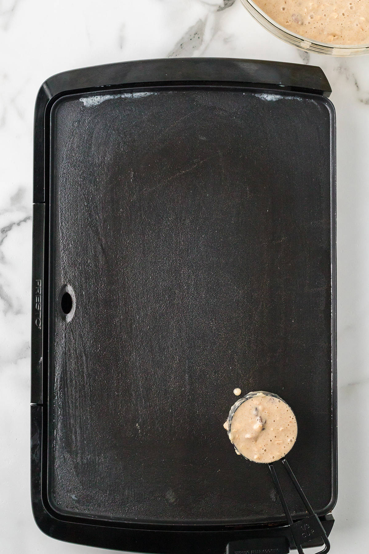 Overhead view of a black griddle with a measuring cup pouring batter onto it, on a white marble counter.