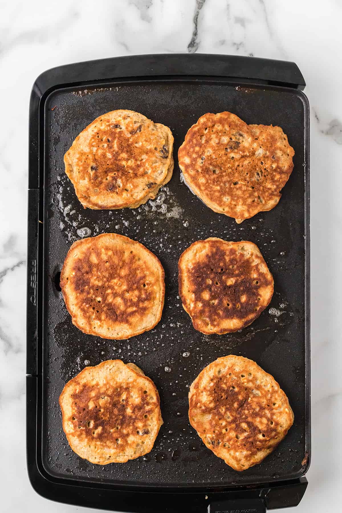 Six golden-brown pancakes cooking on a black electric griddle, seen from above.