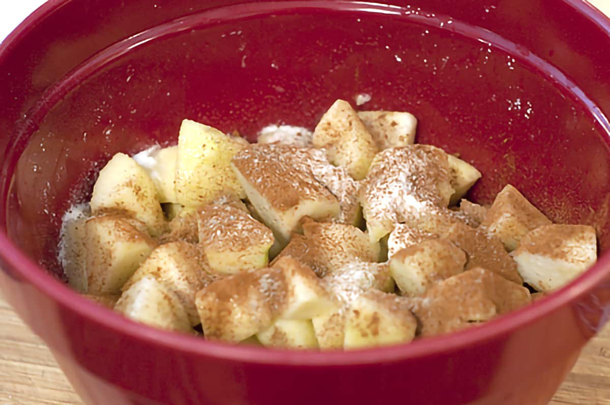 Prepared apples in a red mixing bowl with  lemon juice, cornstarch, sugar, cardamom, cinnamon and orange liqueur.
