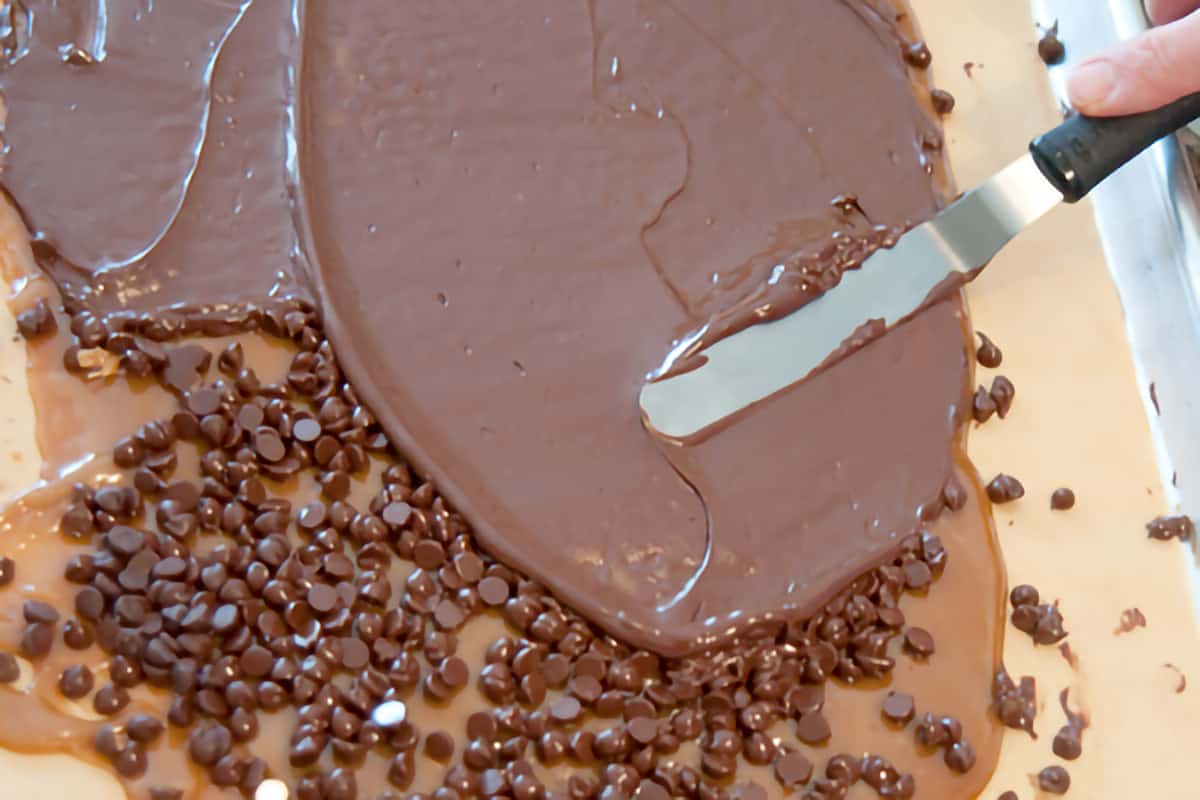 Spreading softened chocolate chips over the toffee layer on a baking sheet.