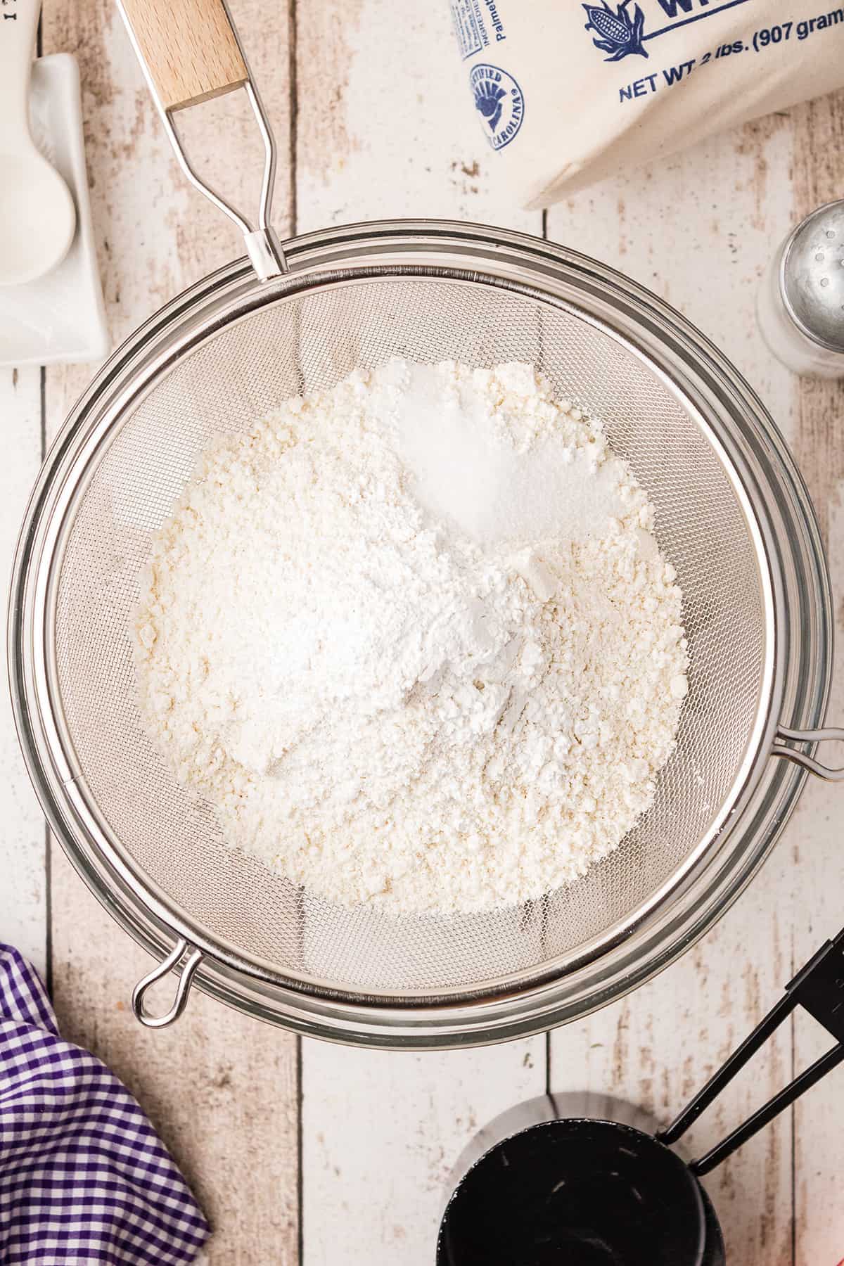 Dry ingredients for the recipe in a sifter over a mixing bowl.