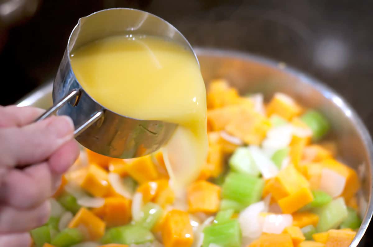 Orange juice in a measuring cup being poured into vegetables in a skillet.