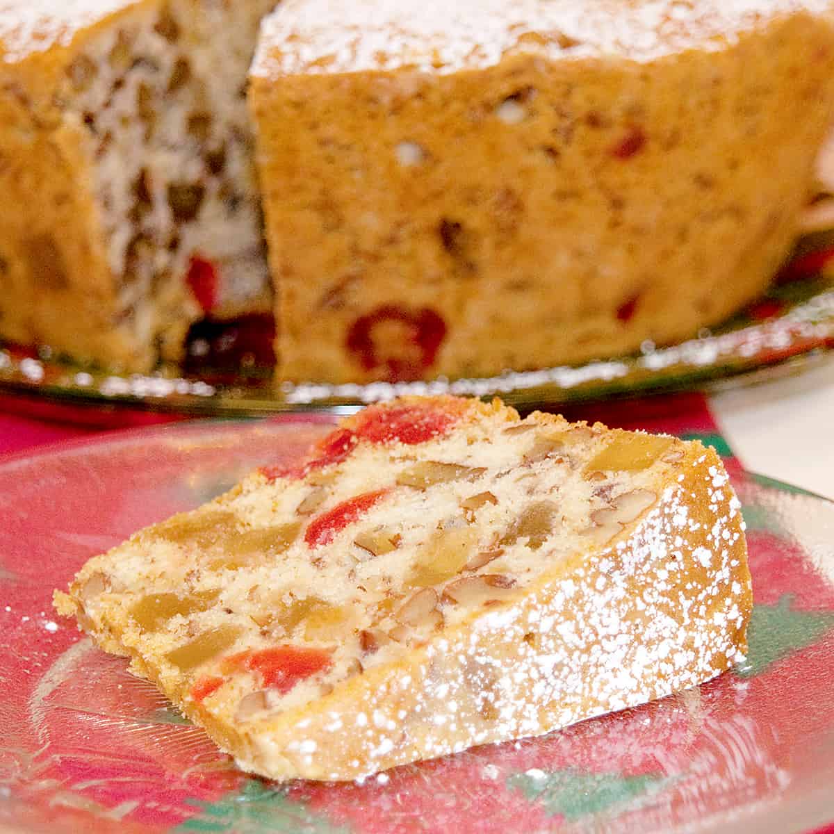 A slice of candied holiday fruitcake on a plate with remaining cake in background.
