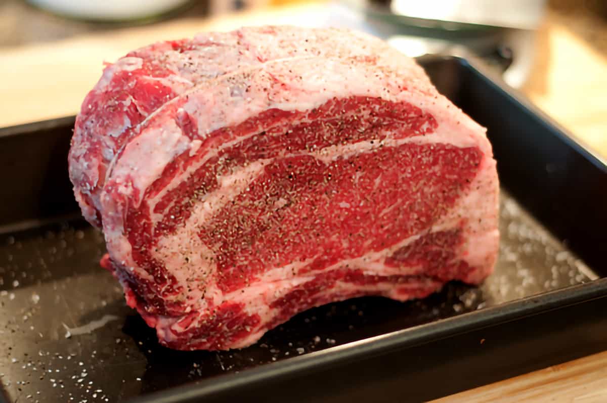 Prime rib generously sprinkled with salt and pepper sitting in a roasting pan.