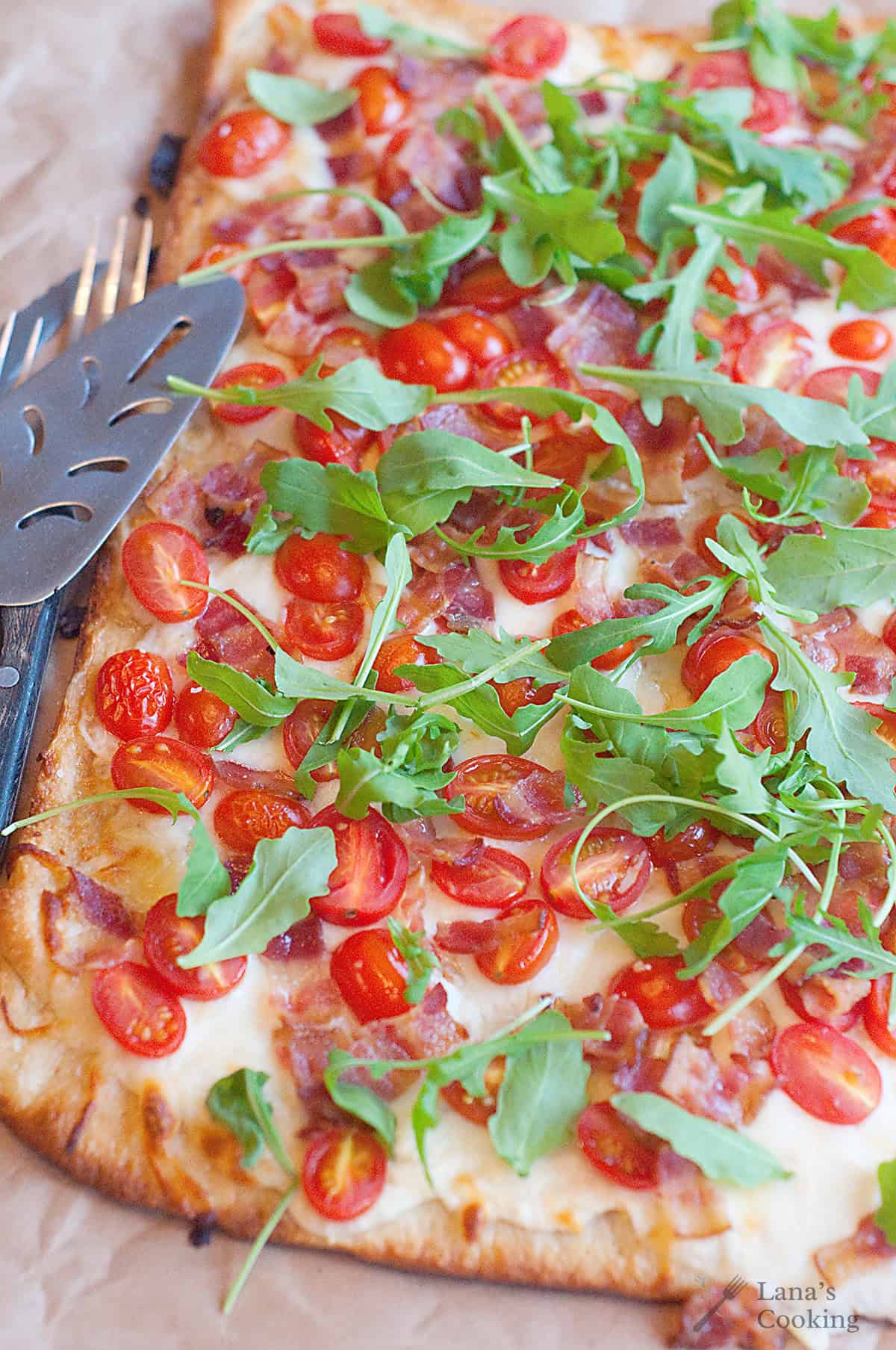 Rectangular flatbread pizza topped with melted cheese, bacon pieces, cherry tomato halves, and fresh arugula, on parchment paper with a metal spatula on the side.