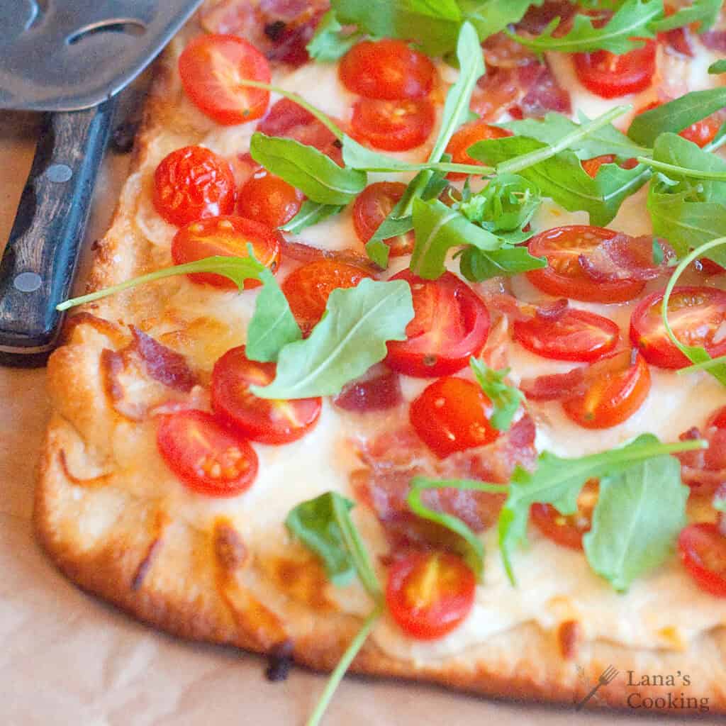 Rectangular flatbread pizza topped with melted cheese, cherry tomatoes, bacon pieces, and fresh arugula leaves on a parchment-lined surface.