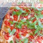 Rectangular BLT pizza topped with bacon, cherry tomatoes, and arugula on a wooden board, next to a metal spatula. Text overlay reads BLT Pizza Under 30 Minutes.