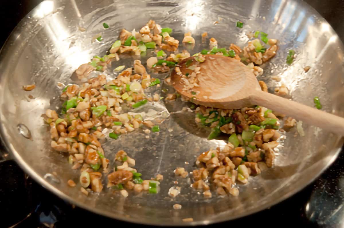 Mushroom stems, green onions, and pecans in a skillet with a wooden spoon.