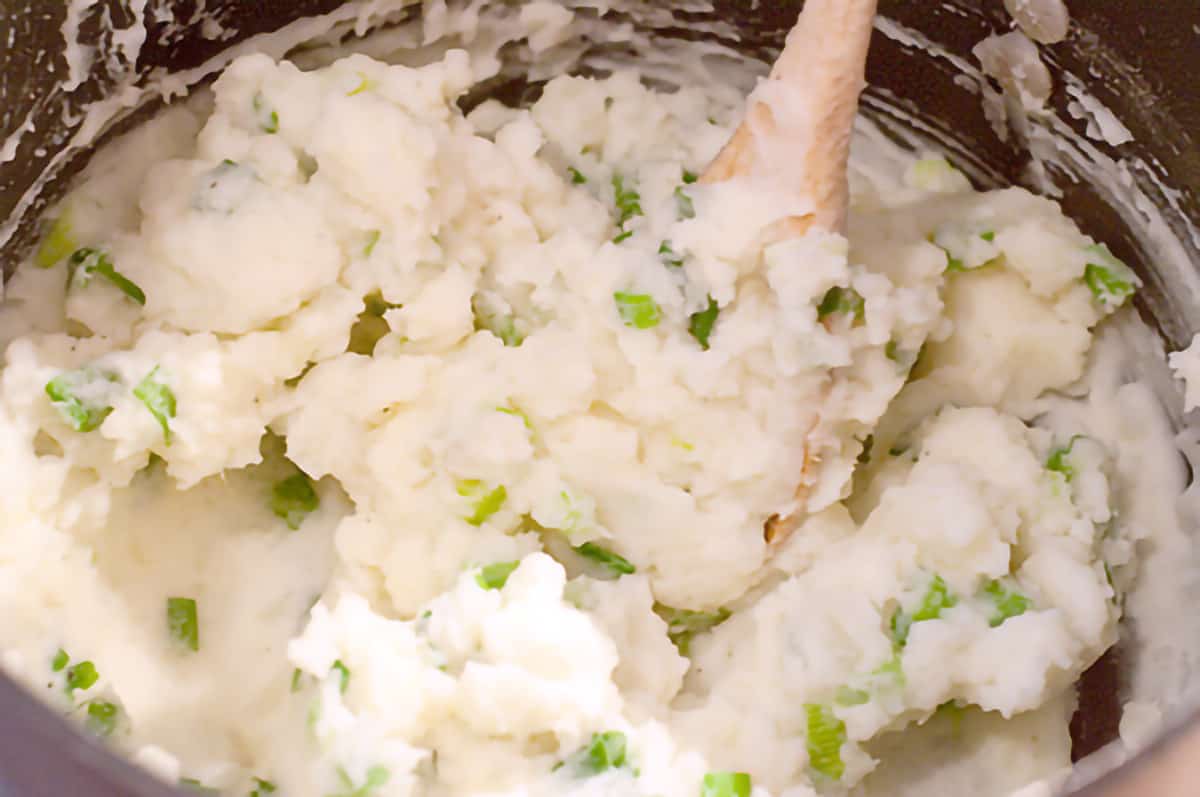 Potatoes, scallions, and milk being mixed with a wooden spoon.