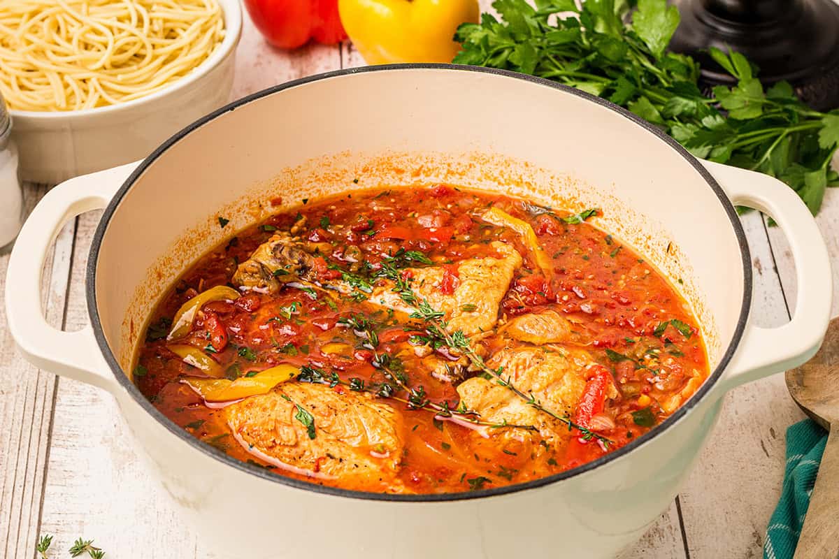 Chicken cacciatore braised in tomato sauce with herbs and vegetables in a white Dutch oven, with pasta and herbs nearby.