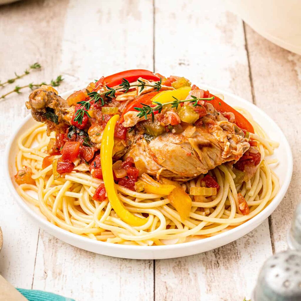 Dutch Oven Chicken Cacciatore over pasta in a white serving bowl on a rustic wooden background.