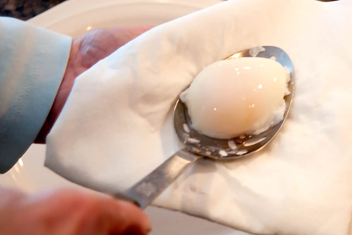 A poached egg in a slotted spoon being blotted on a kitchen towel.