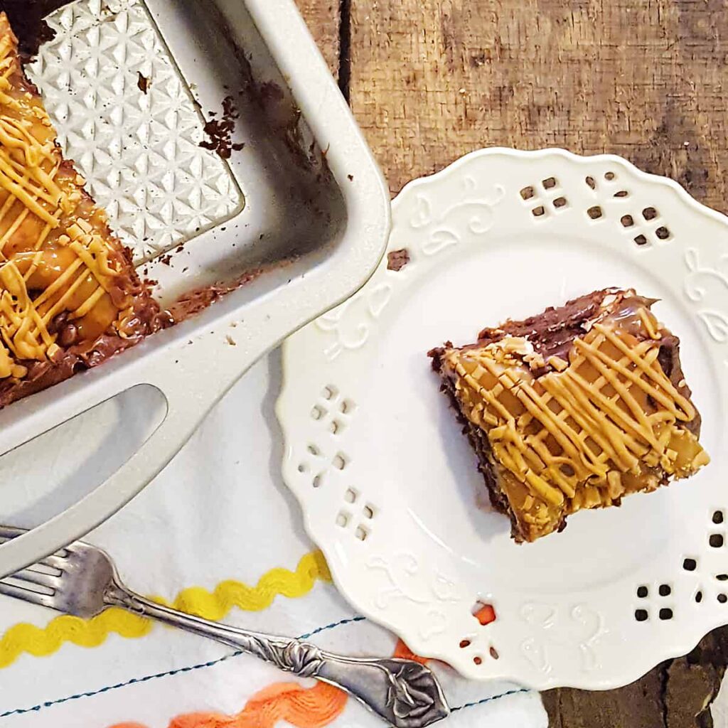 A serving of Easy Caramel Pretzel Brownies on a white serving plate with the full pan of brownies in the background.