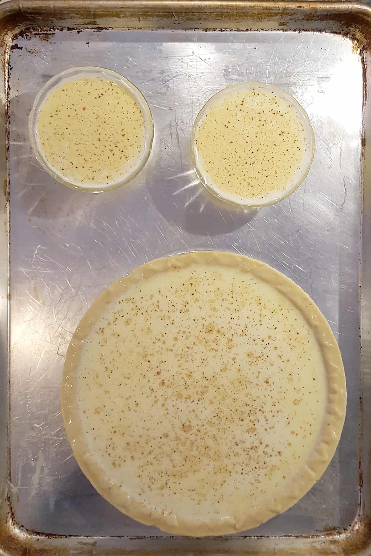 Filled pie crust and two filled custard cups on a baking sheet.