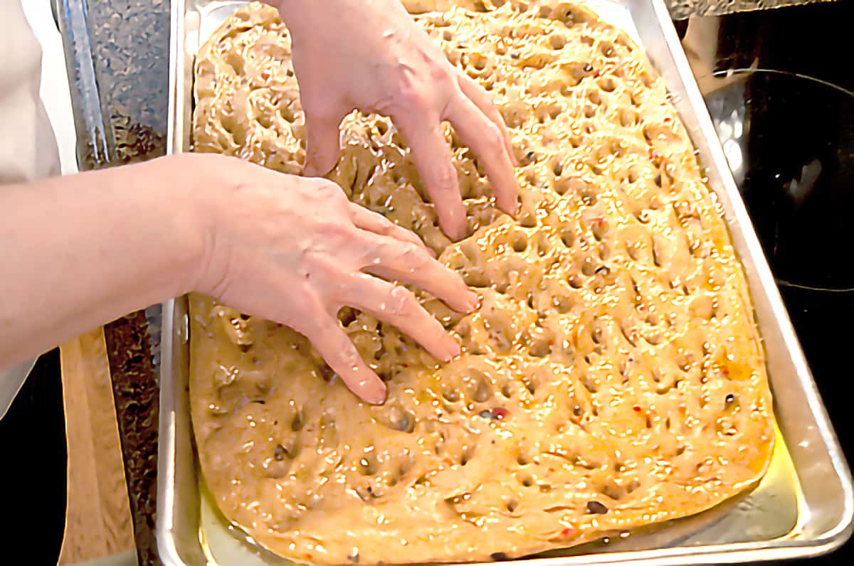 Focaccia dough on a baking sheet with the baker's hands making dimples in the dough.