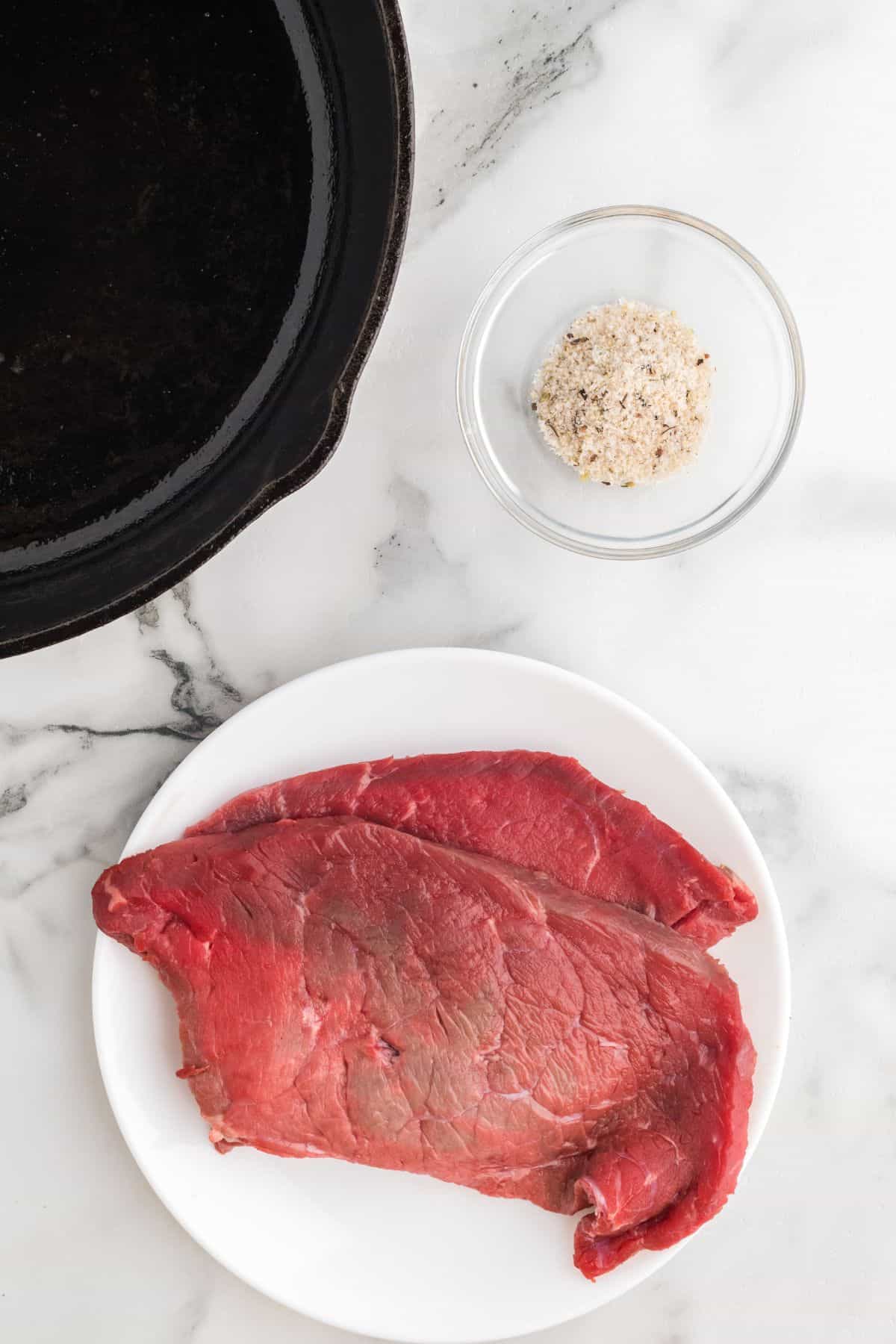 Steak on a white plate pictured with a dish of seasoning next to it.