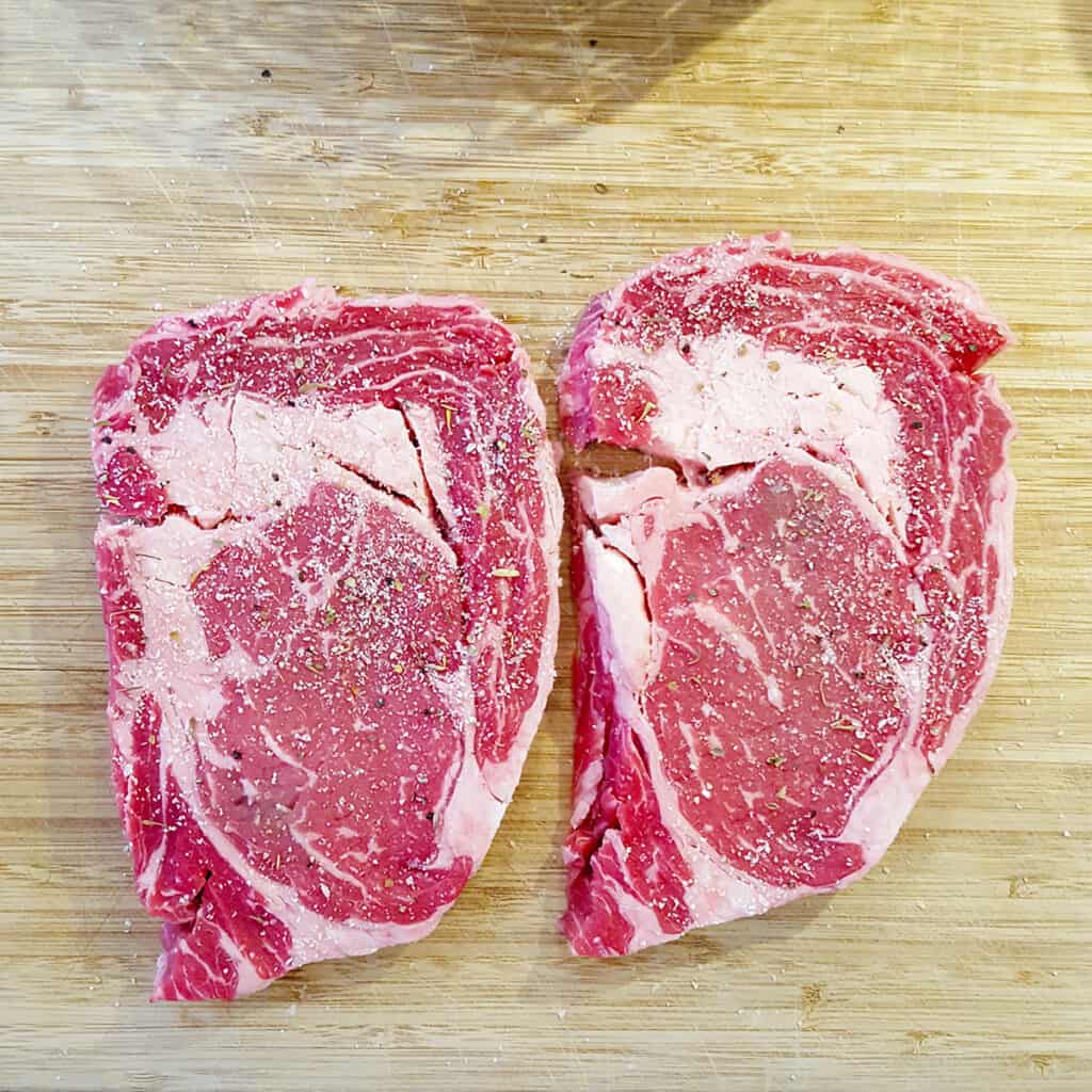 Two thinly cut ribeye steaks on a cutting board sprinkled with seasoning salt.