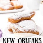 A serving of beignets topped with powdered sugar with a cafe au lait in the background. Text overlay for pinning.