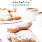 A serving of beignets topped with powdered sugar with a cafe au lait in the background.
