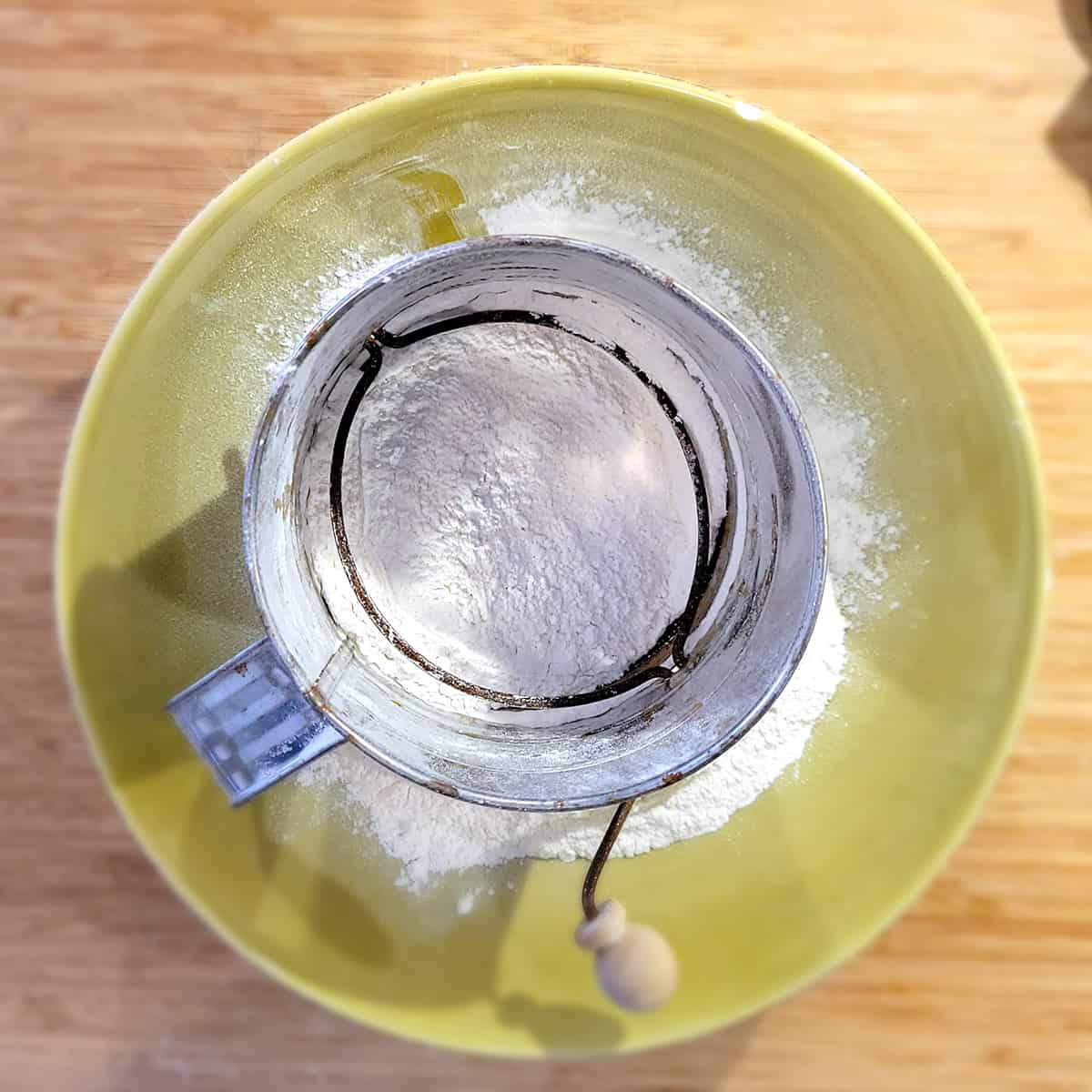 A metal flour sifter sits inside a yellow bowl, sifting white flour onto a wooden surface.