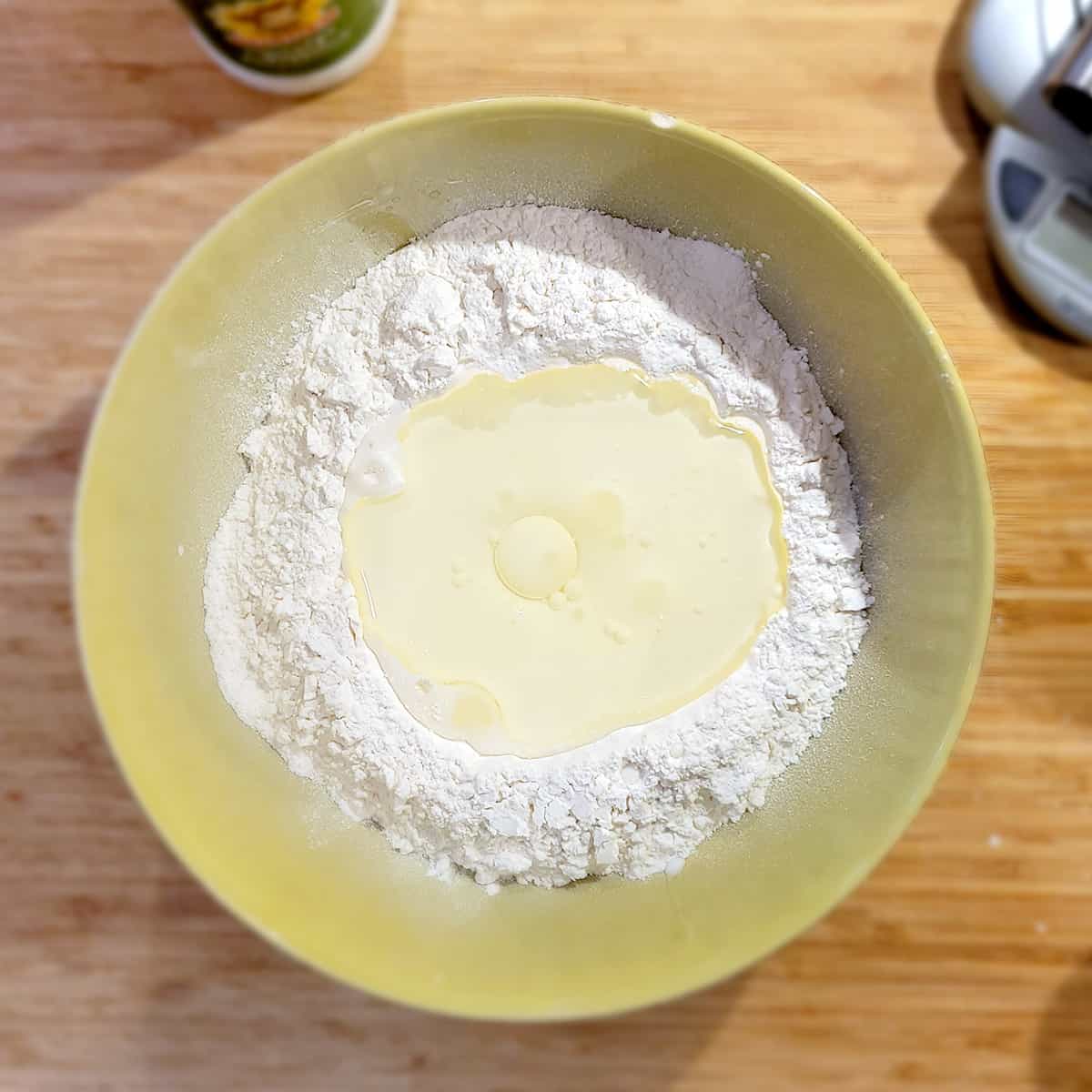 A bowl of flour with a well in the center filled with buttermilk and oil, on a wooden surface.