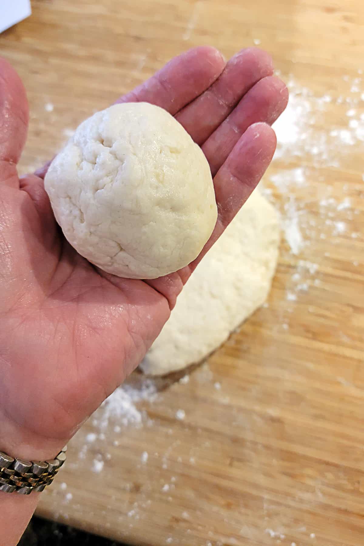 A hand holds a ball of dough above a floured wooden surface with more dough in the background.