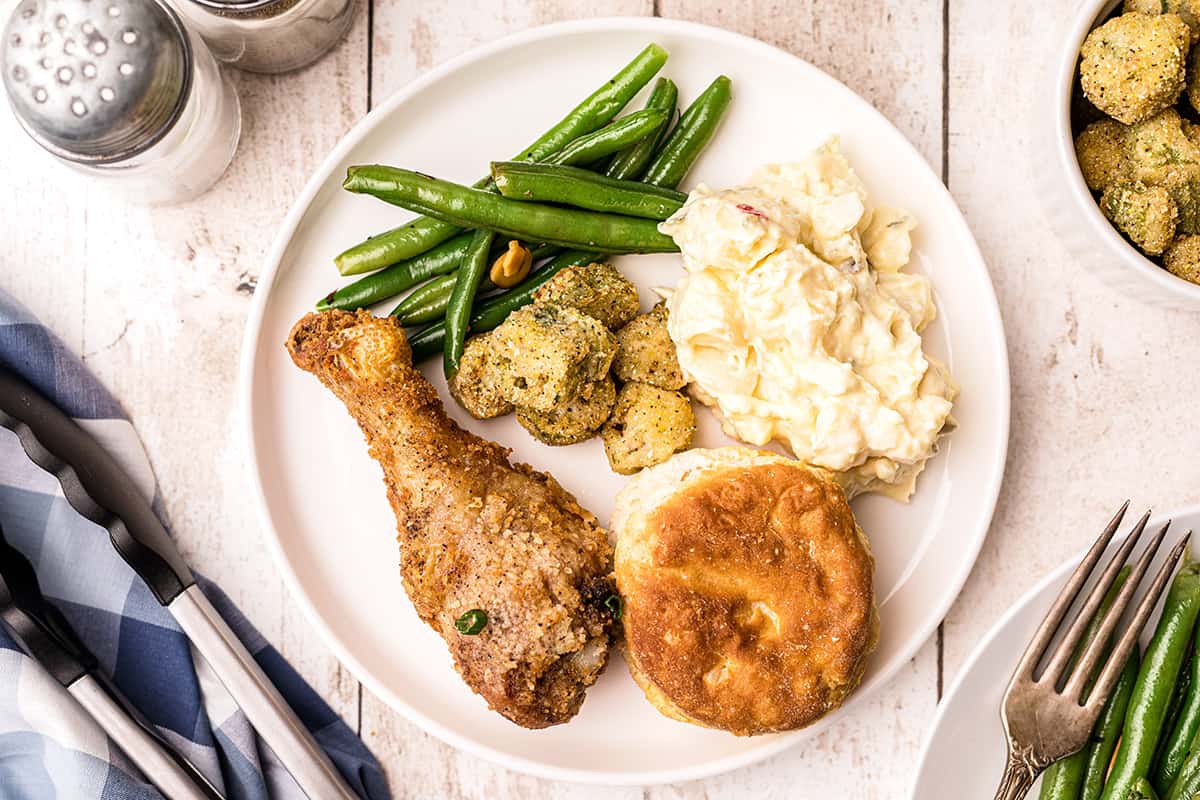 Fried chicken leg on a white dinner plate with green beans, okra, potatoes, and biscuits.