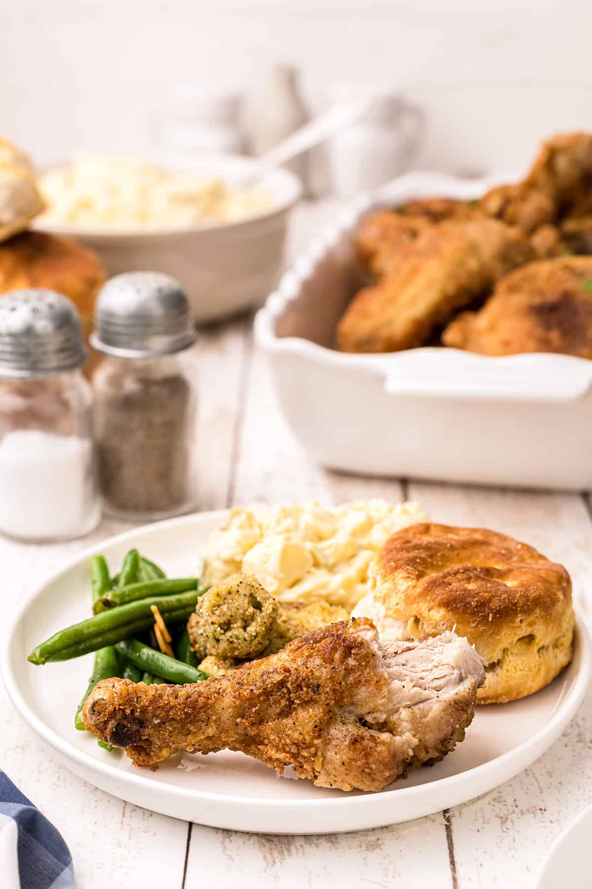 Fried chicken leg on a white dinner plate with green beans, okra, potatoes, and biscuits.