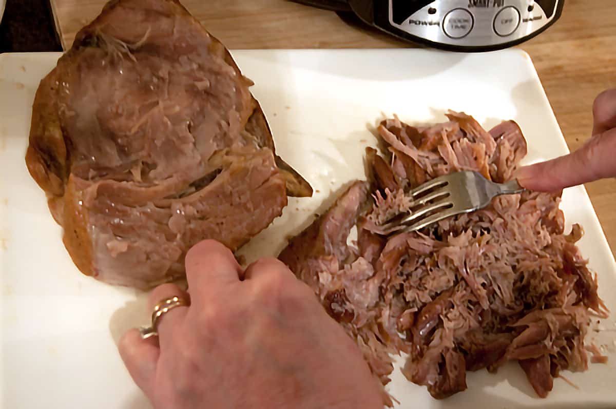 Cooked pork roast being pulled into shreds on a cutting board.
