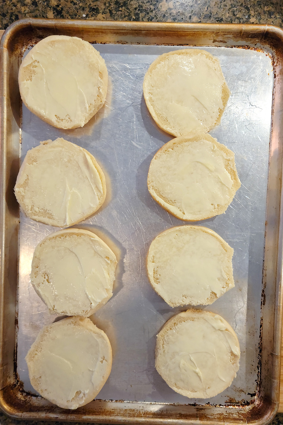 Hamburger buns spread with butter on a baking sheet.