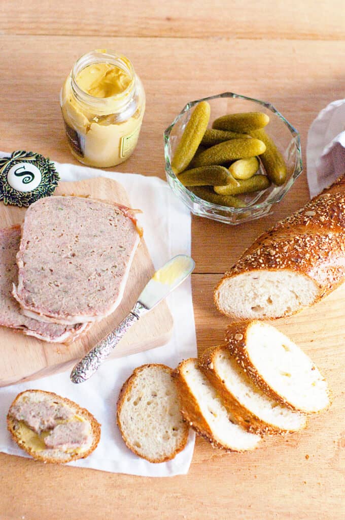 Sliced French country terrine on a serving board with sliced baguette, cornichons, and mustard.