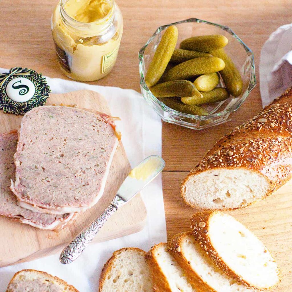 Sliced French country terrine on a serving board with sliced baguette, cornichons, and mustard.