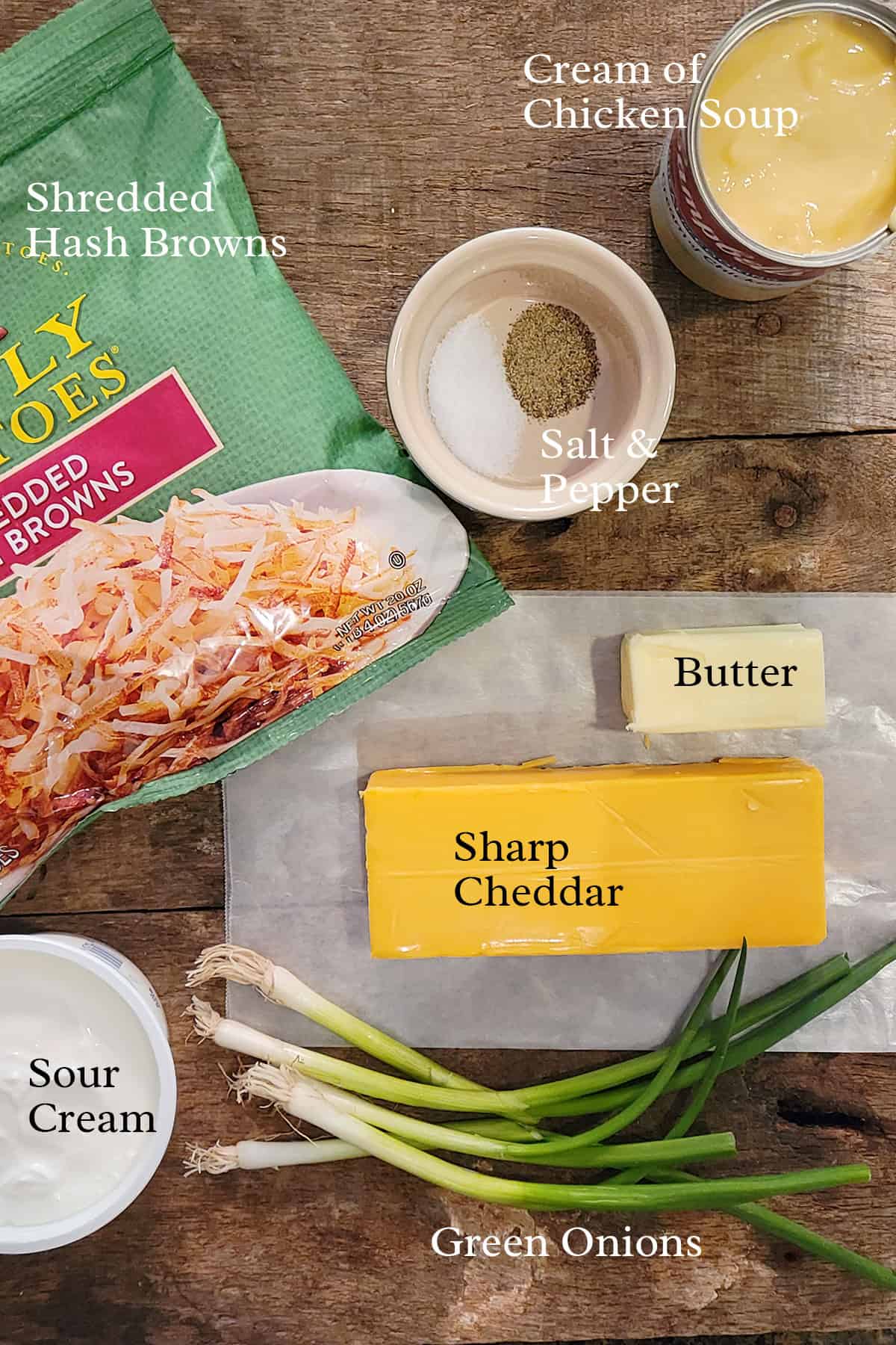 Shredded hash browns, cheddar, sour cream, green onions, butter, soup, salt, and pepper on a wooden table.