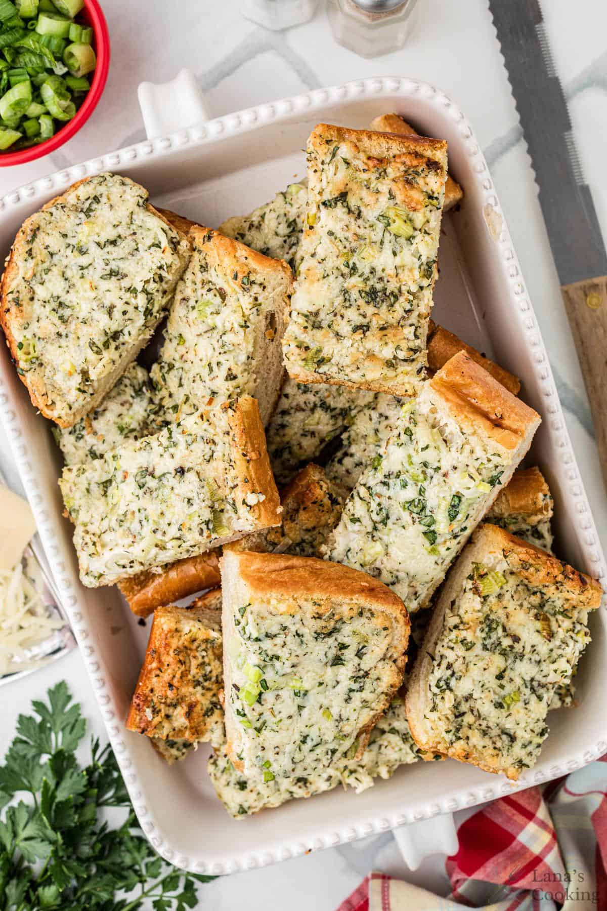 A white dish filled with several slices of garlic bread topped with herbs and cheese. A red bowl with chopped green onions, a knife, and a checkered cloth are visible nearby.
