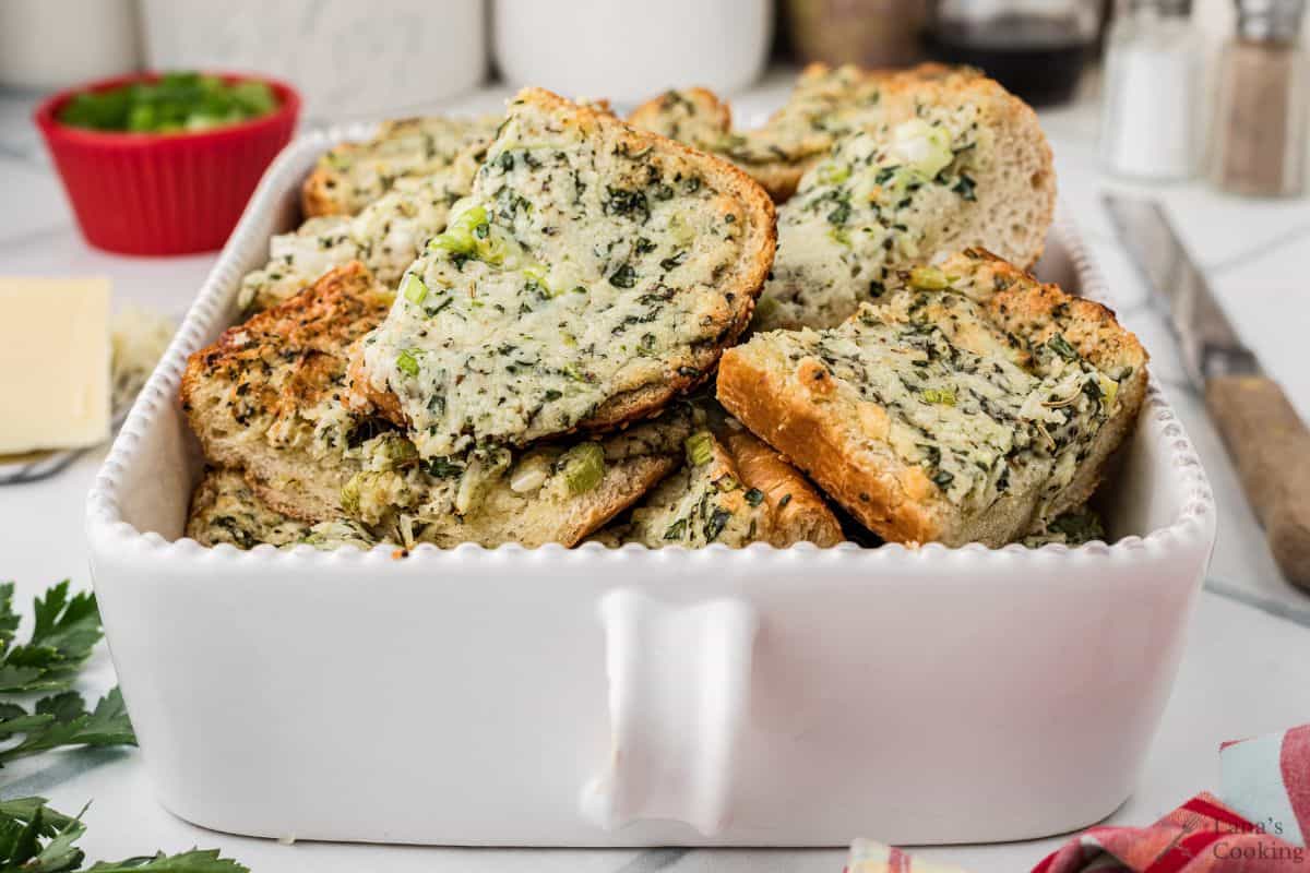 A white rectangular dish filled with slices of bread topped with a green spinach and cheese mixture, set on a kitchen counter with herbs and ingredients in the background.