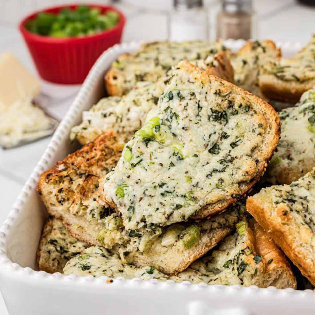 A white dish filled with slices of bread topped with a mixture of melted cheese, chopped spinach, and green onions. In the background, there are salt and pepper shakers and a red bowl filled with chopped green onions.