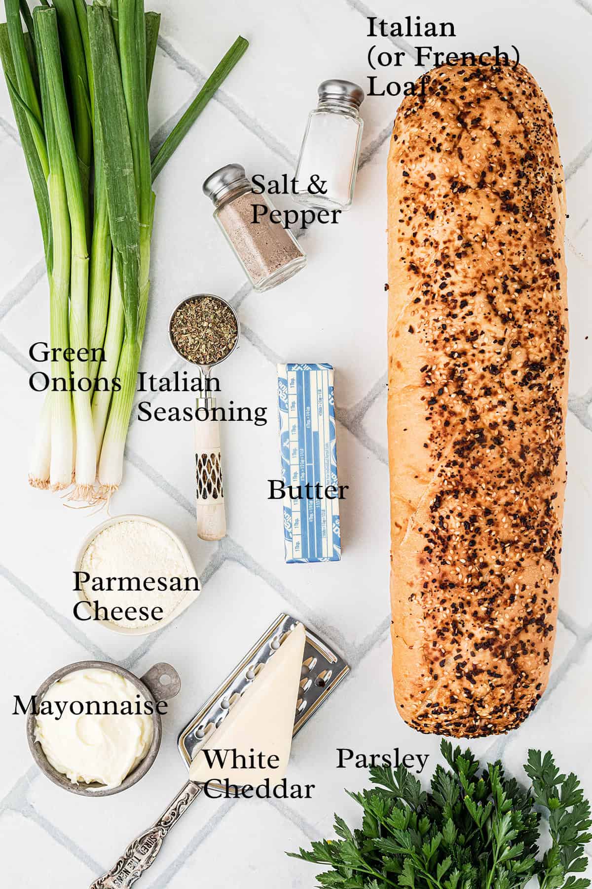 A loaf of Italian bread, green onions, salt, pepper, Italian seasoning, butter, parsley, mayonnaise, Parmesan cheese, and a wedge of white cheddar are arranged on a white tiled surface.
