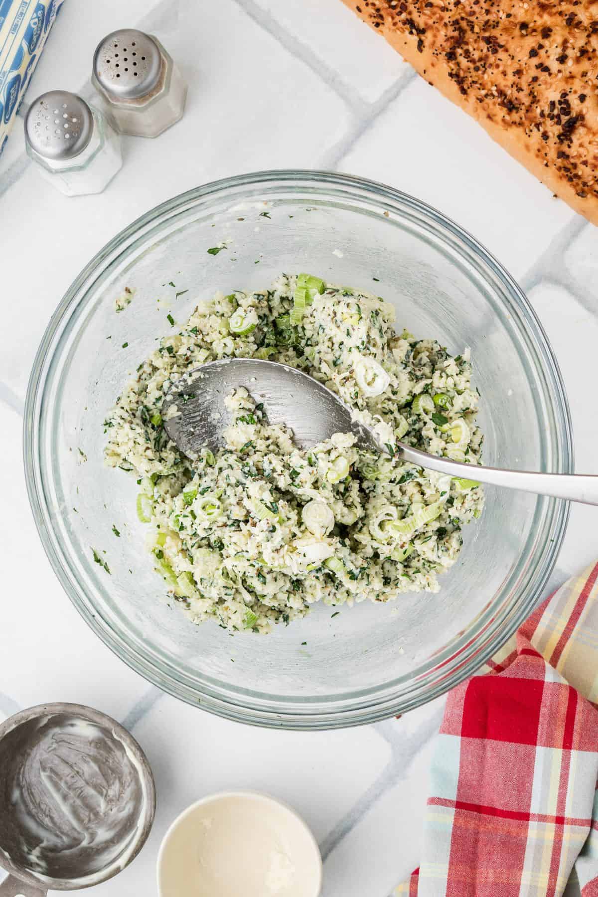 A glass mixing bowl contains a mixture of chopped green herbs, scallions, and ricotta cheese being stirred with a metal spoon. Nearby are a salt and pepper shaker, a piece of bread, and a red plaid cloth.