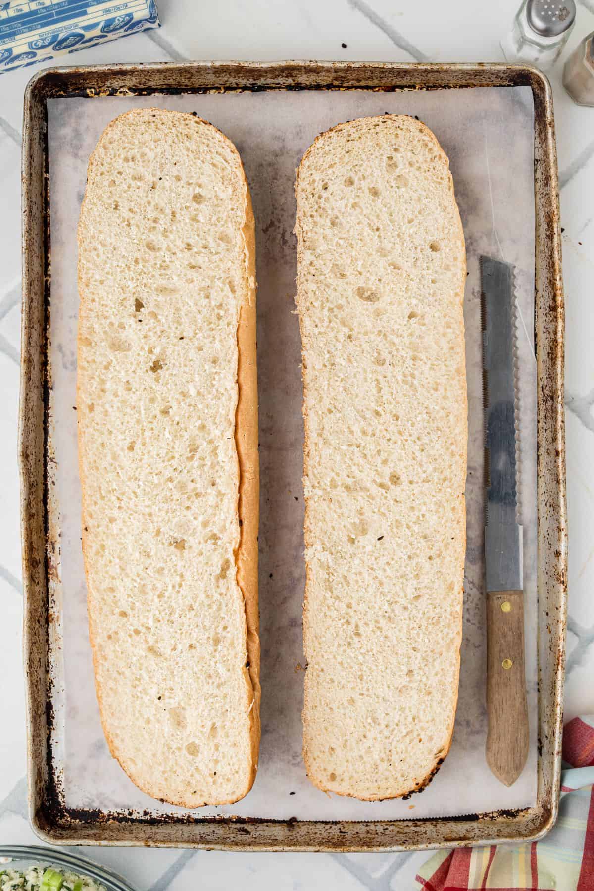 A baking sheet lined with parchment paper holds a loaf of bread sliced in half lengthwise. A bread knife with a wooden handle is placed next to the bread on the right side of the tray.