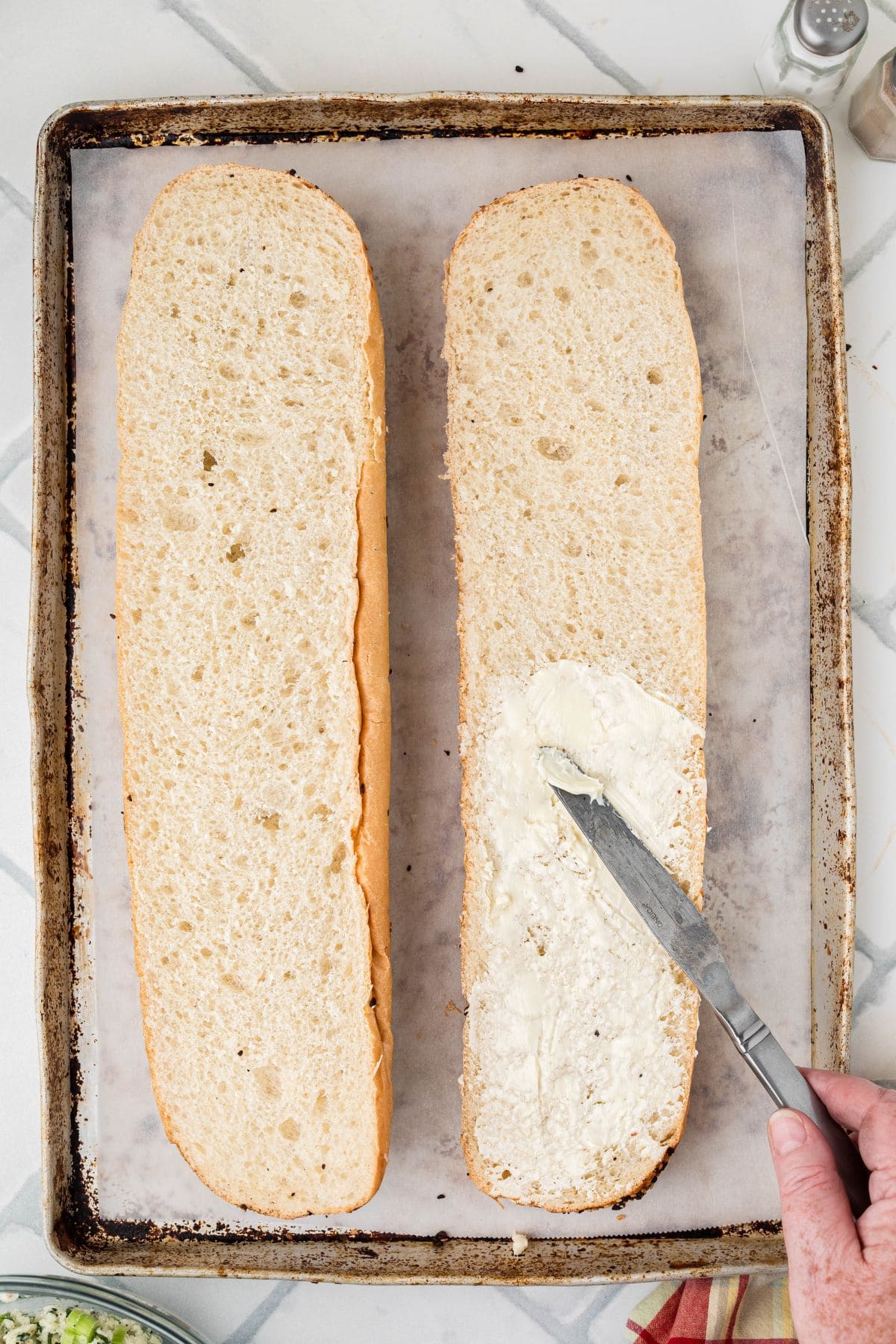 A hand spreads butter on one half of a sliced baguette using a butter knife. The bread halves are on a parchment-lined baking sheet.