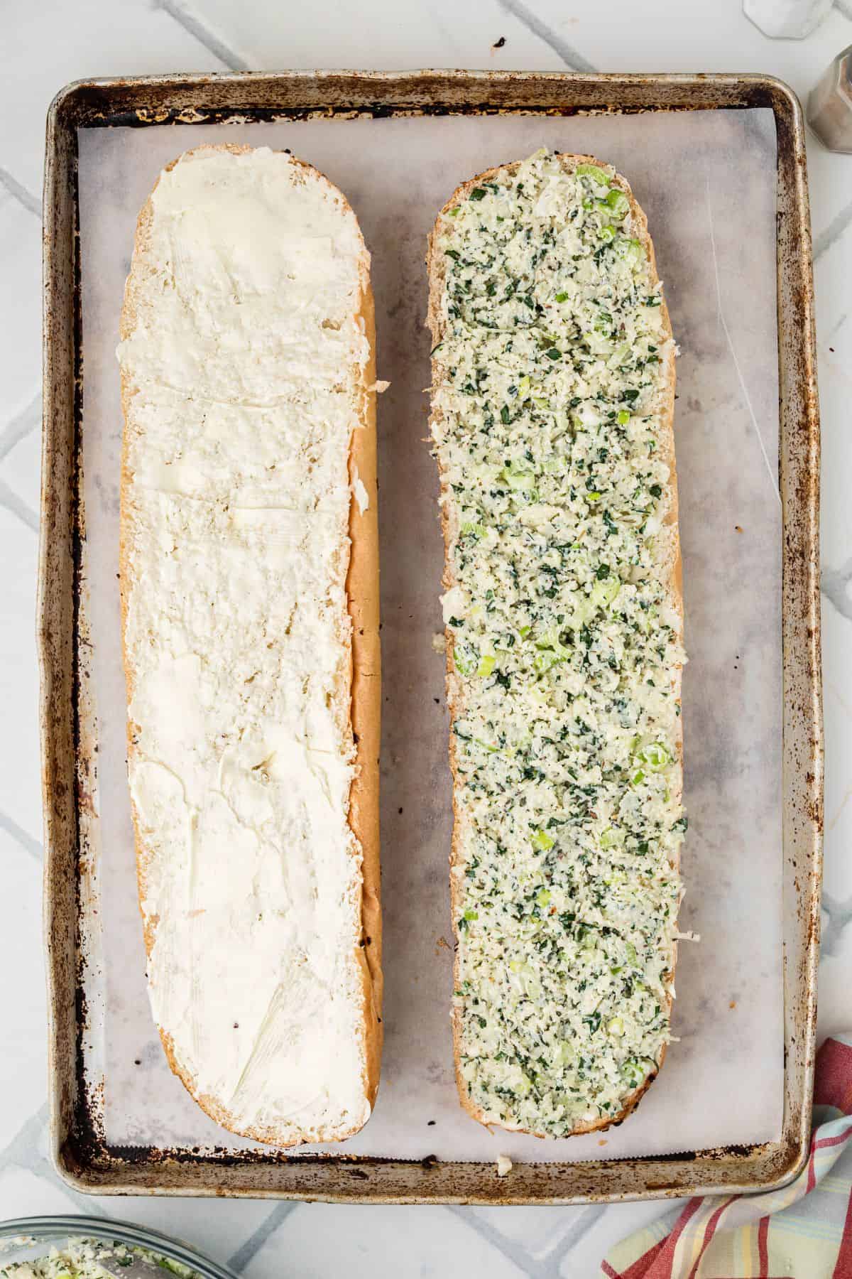 A baking sheet holds a split loaf of bread. One half is spread with a white creamy layer, and the other half is topped with a chunky spinach and cheese mixture. Both halves are placed side by side, cut side up.
